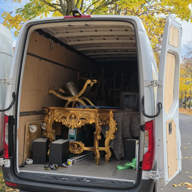 Offene Rückseite eines weißen Lieferwagens, der neben einem Baum mit gelben Herbstblättern geparkt ist, mit Blick auf verzierte goldene Möbel und verschiedene Gegenstände im Inneren.