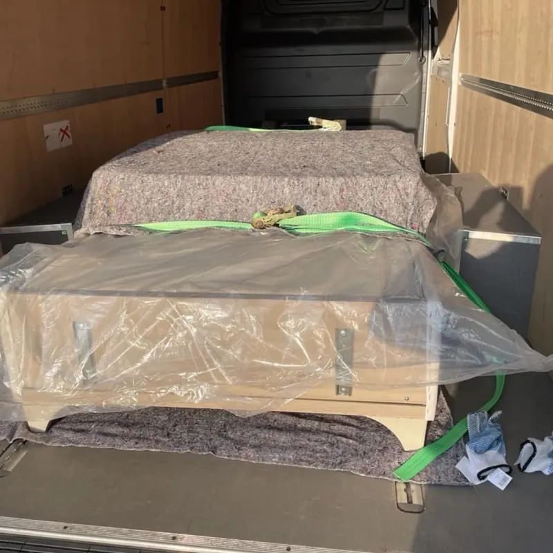 Holzmöbelstück, in Plastikfolie eingewickelt und mit grünen Gurten gesichert, in einem Transportwagen.