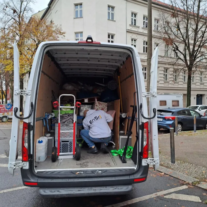 Eine Person, die einen EnSa-Hoodie trug, kauerte im hinteren Teil eines offenen weißen Lieferwagens, der mit verschiedenen Umzugsgegenständen und Ausrüstung gefüllt war.